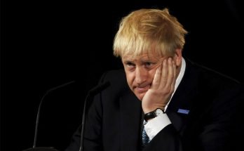 A forlorn looking Boris Johnson sits in front of a microphone against a black background