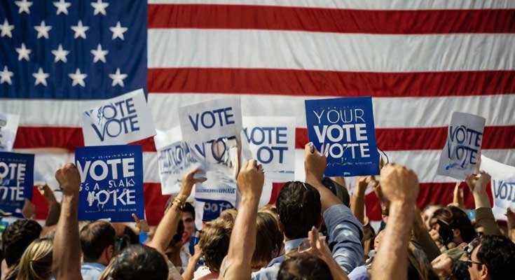 People holding up signs saying "Vote" in front of a US flag